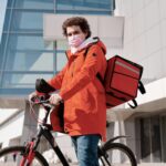 An urban cyclist wearing a face mask and carrying a thermal bag for food delivery, outdoors.
